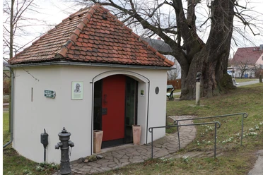 Small building with a red roof and red door, surrounded by trees and a sidewalk; next to it a fire hydrant and a bench on a meadow.