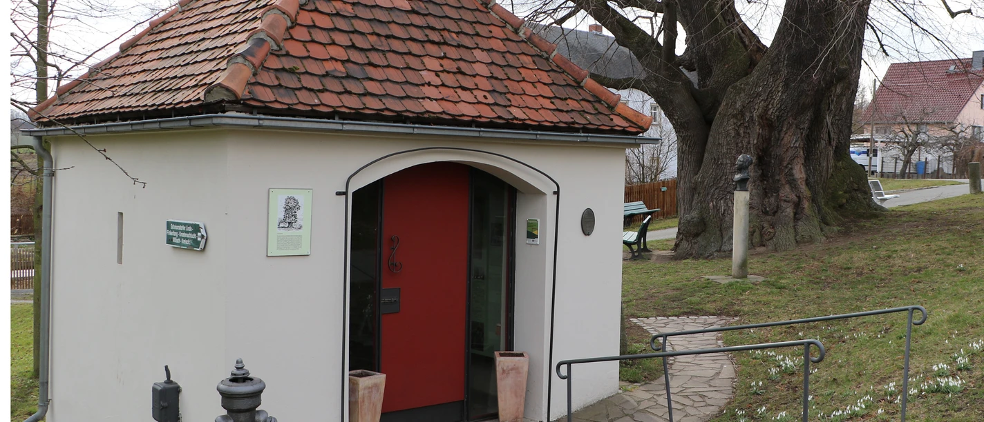 Lindenmuseum "Clara Schumann" Kleines Gebäude mit rotem Dach und roter Tür, umgeben von Bäumen und einem Gehweg; daneben ein Hydrant und eine Bank auf einer Wiese.