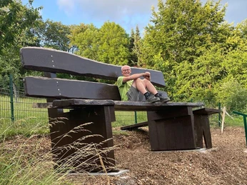 Riesenbank im Ellerbachtal Eine übergroße Holzbank im Grünen mit einem Mann, der entspannt darauf sitzt.