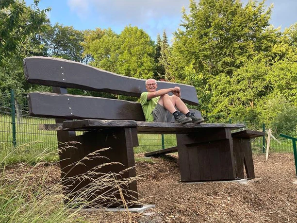 Riesenbank im Ellerbachtal Eine übergroße Holzbank im Grünen mit einem Mann, der entspannt darauf sitzt.