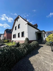 Zweistöckiges Haus mit Satteldach, weißer Fassade, von Hecken umgeben, sonniger Himmel im Hintergrund.