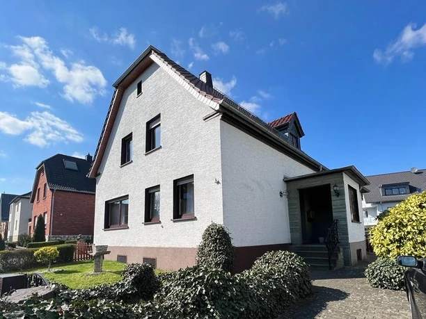 Fewo Wewer, Aussenansicht Zweistöckiges Haus mit Satteldach, weißer Fassade, von Hecken umgeben, sonniger Himmel im Hintergrund.