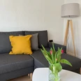 Graues Ecksofa mit gelbem Kissen, daneben Stehlampe. Im Vordergrund Tulpenvase auf Couchtisch.