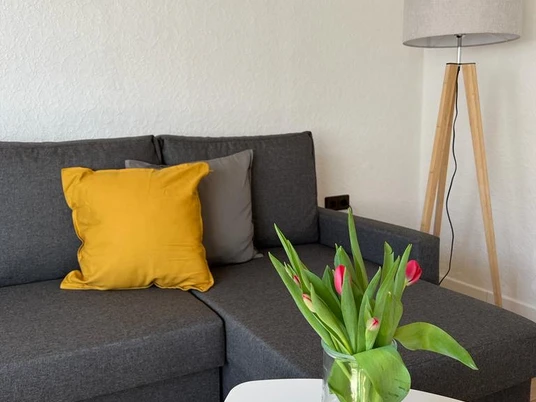 Graues Ecksofa mit gelbem Kissen, daneben Stehlampe. Im Vordergrund Tulpenvase auf Couchtisch.
