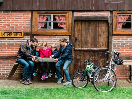 Eine Familie sitzt vor einem Backhaus auf einer Bank, betrachtet eine Landkarte; zwei Fahrräder stehen daneben.