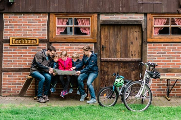 Thorsten-Hennig-Fotografie-Fahrradtouren-Hövelhof-2019-A9_05074.jpg Eine Familie sitzt vor einem Backhaus auf einer Bank, betrachtet eine Landkarte; zwei Fahrräder stehen daneben.