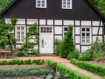 Fachwerkhaus mit Pflanzendeko, Garten am Vordergrund, einladende Terrasse mit bunten Stühlen.