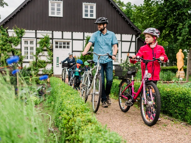 Thorsten-Hennig-Fotografie-Fahrradtouren-Hövelhof-2019-A9_06174.jpg Familie mit Fahrrädern vor einem Fachwerkhaus in ländlicher Umgebung, umgeben von grünen Hecken.