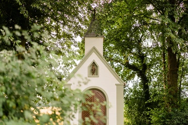 Weiße Kapelle in Waldlichtung, spitze Dachform, umgeben von hohen Bäumen und blühenden Sträuchern.