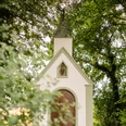 Weiße Kapelle in Waldlichtung, spitze Dachform, umgeben von hohen Bäumen und blühenden Sträuchern.