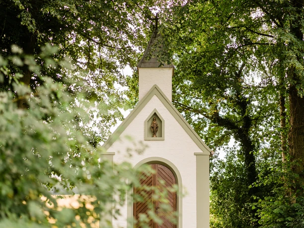 Rodehuths Kapelle Hövelhof-Espeln Weiße Kapelle in Waldlichtung, spitze Dachform, umgeben von hohen Bäumen und blühenden Sträuchern.