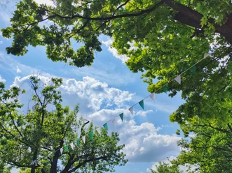 Bunte Wimpel hängen zwischen grünen Bäumen unter einem sonnigen, leicht bewölkten Himmel.