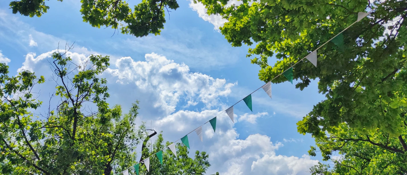 Bunte Wimpel hängen zwischen grünen Bäumen unter einem sonnigen, leicht bewölkten Himmel.