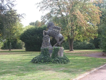 Bronzestatue eines Pferdes mit Reiter auf einem Sockel, umgeben von gepflegtem Rasen und Bäumen.