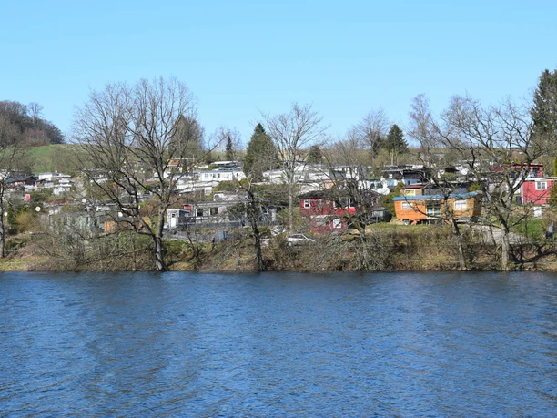 Brucher Talsperre Wasserfläche mit Freizeitgrundstücken und Wohnmobilen entlang eines bewaldeten Ufers unter blauem Himmel.