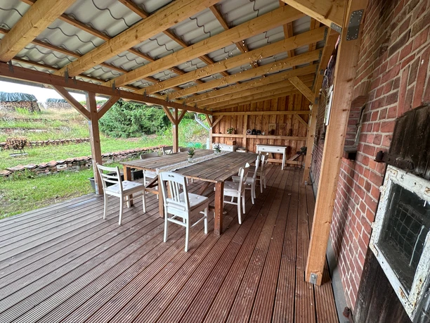 lebensrau-kräuter-amelinghausen1.JPG Überdachte Holzterrasse mit langem Holztisch und weißen Stühlen an einem Backsteingebäude.