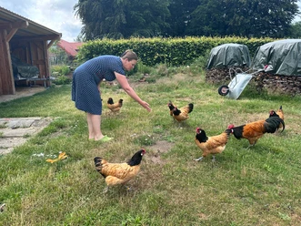 Frau im gepunkteten Kleid füttert braune Hühner auf einer grünen Wiese.