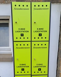 Vier leuchtend grüne E-Bike Ladesäulen der Marke Strandterrassen vor einer verputzten Wand.