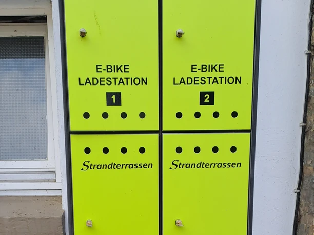 Vier leuchtend grüne E-Bike Ladesäulen der Marke Strandterrassen vor einer verputzten Wand.