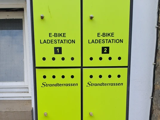 E-Ladestation an den Strandterrassen Vier leuchtend grüne E-Bike Ladesäulen der Marke Strandterrassen vor einer verputzten Wand.