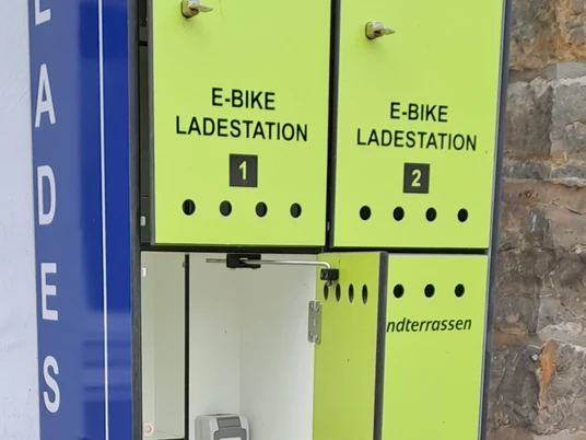 E-Ladestation an den Strandterrassen geöffnet Ein E-Bike-Ladestation mit vier Schließfächern, integriert in eine Steinwand, bietet praktischen Zugang.