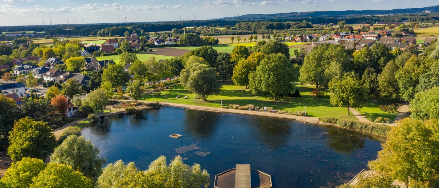 Uitzicht op een bebost meer met een steiger in het midden van een groen landschap en verspreide huizen.