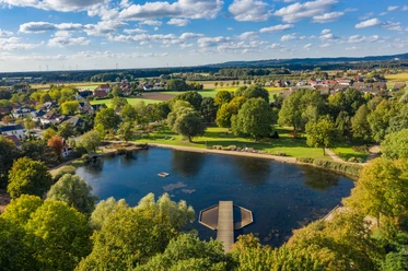 Der Kurpark in Bad Laer aus der Vogelperspektive.jpg Uitzicht op een bebost meer met een steiger in het midden van een groen landschap en verspreide huizen.