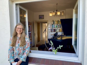 Fenstermusik Eine Frau mit langem, blondem Haar lehnt lächelnd an einem geöffneten Fenster, im Hintergrund ein Klavier.