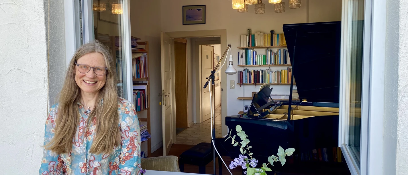 Fenstermusik Eine Frau mit langem, blondem Haar lehnt lächelnd an einem geöffneten Fenster, im Hintergrund ein Klavier.