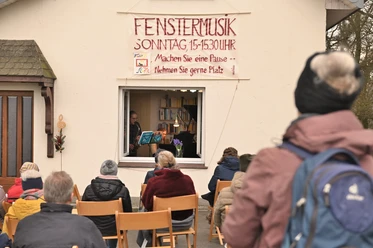 Fenstermusik Personen sitzen im Freien und lauschen einem Musiker, der aus einem Fenster heraus spielt.