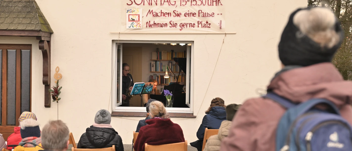Fenstermusik Personen sitzen im Freien und lauschen einem Musiker, der aus einem Fenster heraus spielt.