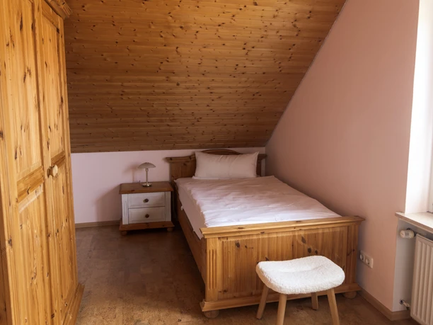 Schlafzimmer mit Einzelbett unter Dachschräge, aus Holz gefertigt, mit Nachttisch und Hocker.