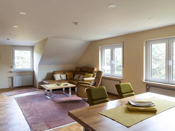 Termuende1Wohn-Esszimmer Sitzecke.jpg Modernes Wohn-Esszimmer mit gemütlicher Sitzecke, Holzmöbeln und großen Fenstern mit Gartenblick.