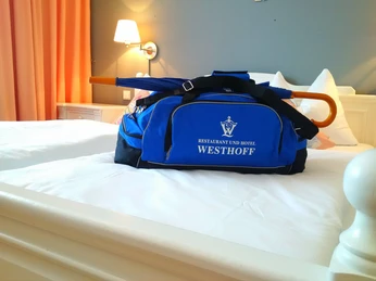 Hotel Westhoff Tasche_CCBYSA_Teutoburger Wald_Stadt_Schloss_Holte-Stukenbrock_Hotel Westhoff.jpg Eine blaue Reisetasche mit dem Schriftzug "Restaurant und Hotel Westhoff" liegt auf einem Doppelbett.