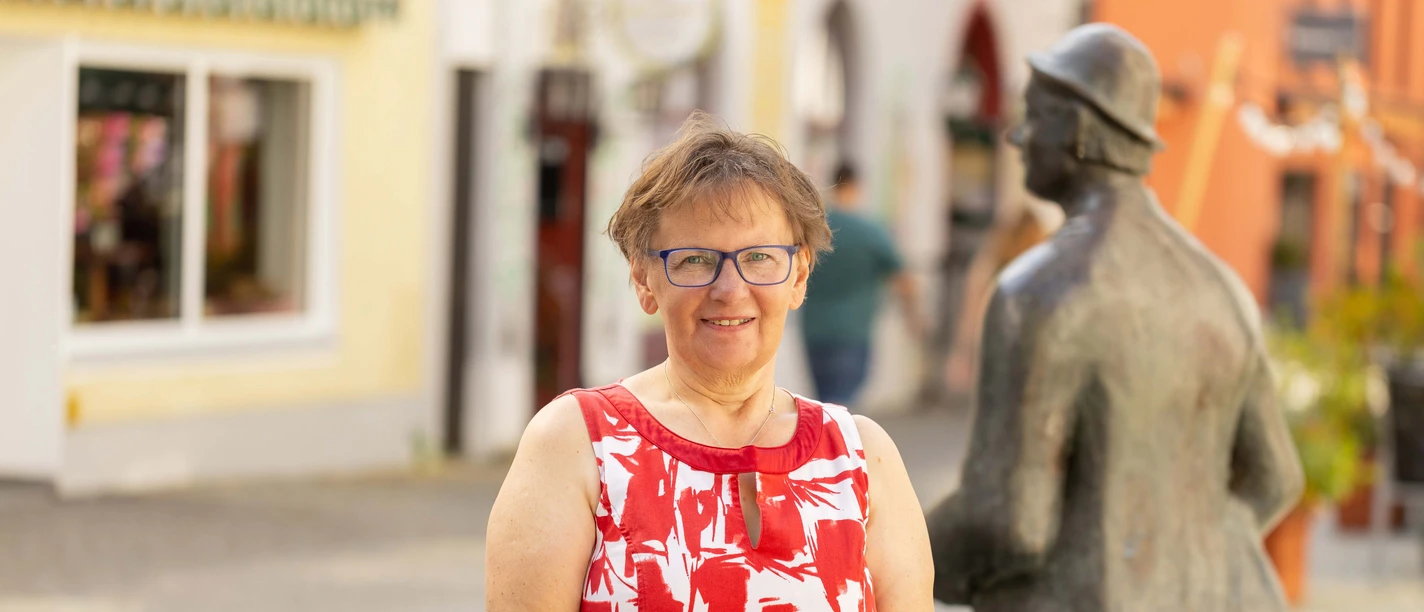 Faulenzer & Fürkäufler - Die Brauchtumsführung in Günzburg Ältere, lächelnde Frau in rotem Kleid neben einer Bronzeskulptur auf einem sonnigen Platz.