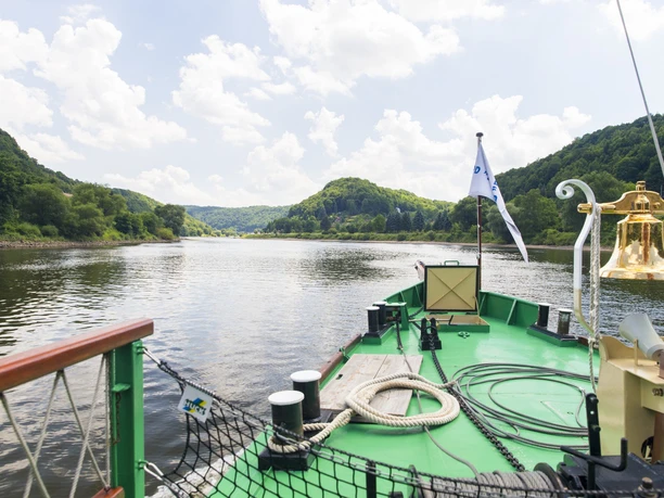 View from the deck of a steamboat onto a calm river, surrounded by wooded hills under a slightly cloudy sky.