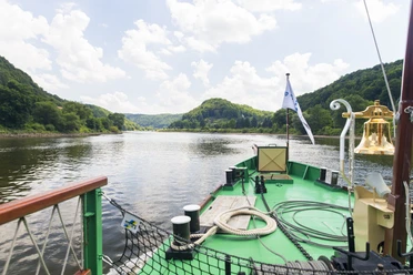 Sächsische Dampfschifffahrt Blick vom Deck eines Dampfschiffes auf einen ruhigen Fluss, umgeben von bewaldeten Hügeln unter einem leicht bewölkten Himmel.
