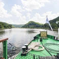 Sächsische Dampfschifffahrt Blick vom Deck eines Dampfschiffes auf einen ruhigen Fluss, umgeben von bewaldeten Hügeln unter einem leicht bewölkten Himmel.