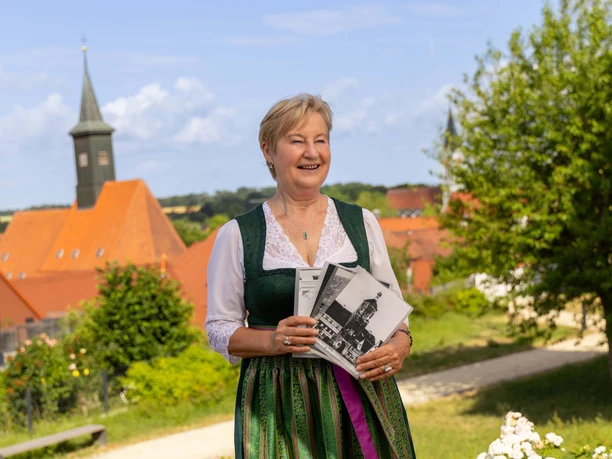 Günzburg und seine Geschichte - Der Klassiker unter den Stadtführungen Auf dem Bild ist eine lächelnde Frau in traditioneller Tracht zu sehen, die vor einem malerischen, historischen Hintergrund mit rotem Ziegeldach steht und Broschüren in der Hand hält.