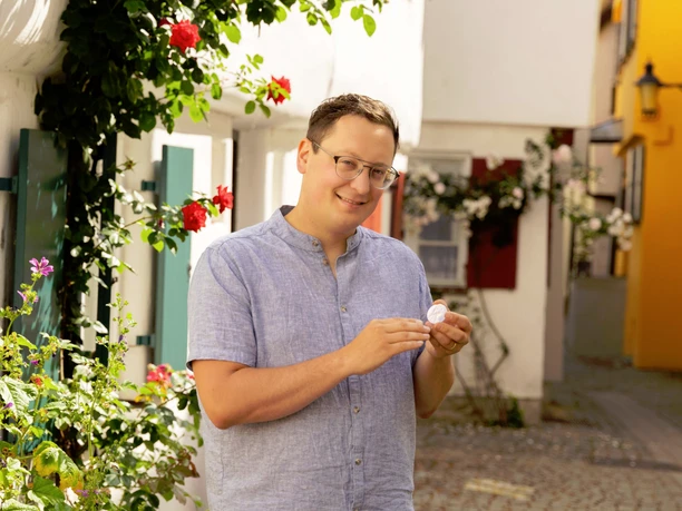 Günzburg und seine Geschichte - Der Klassiker unter den Stadtführungen Ein lächelnder Mann mit Brille und hellem Leinenhemd steht in einer bunten, blumengeschmückten Gasse.