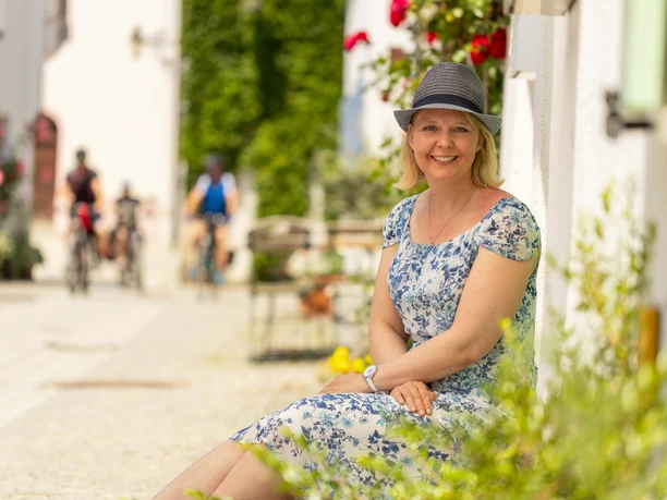 Eine lächelnde Frau in einem blumigen Kleid sitzt in einer sonnigen Gasse von Günzburg, im Hintergrund Radfahrer.