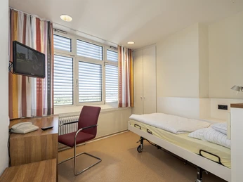 DSCF2504.jpg Ein schlicht eingerichtetes Einzelzimmer mit Krankenhausbett, modernem Fernseher und Fenstern mit Jalousien.