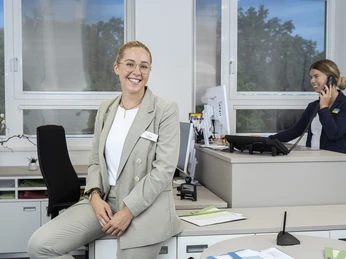 DSCF2580final.jpg Eine lächelnde Frau in Business-Kleidung lehnt an einem Schreibtisch. Dahinter bedient eine weitere Frau einen Computer.