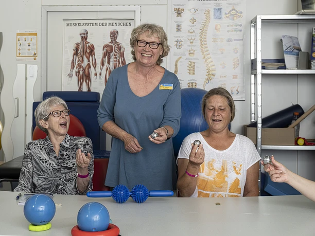 Drei Frauen bei einer Physiotherapie-Übung, lächelnd und mit Therapiegeräten in der Hand.