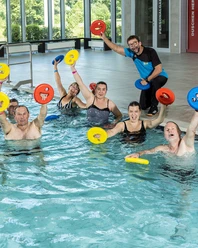 Schwimmbad.jpg Eine Gruppe von schwimmenden Erwachsenen hebt bunte Wasserhanteln lachend im Innenpool.
