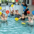 Schwimmbad.jpg Eine Gruppe von schwimmenden Erwachsenen hebt bunte Wasserhanteln lachend im Innenpool.