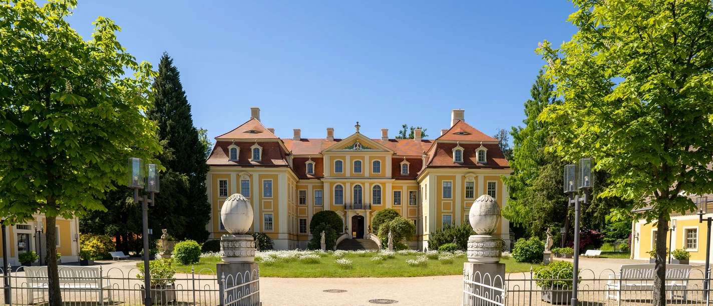 Barockschloss-Rammenau-Foto-Ben-Walther_001-klein.jpg Barockschloss Rammenau im Sommer, gelb mit roten Dächern, flankiert von Bäumen, blauer Himmel.