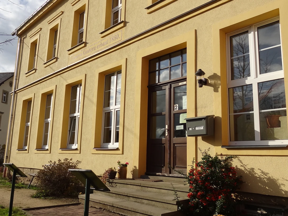 Touristinformation Altendorf Gelbes Gebäude mit großen Fenstern und einer braunen Tür, daneben ein Briefkasten; sonnige Atmosphäre mit blühenden Sträuchern im Vordergrund.