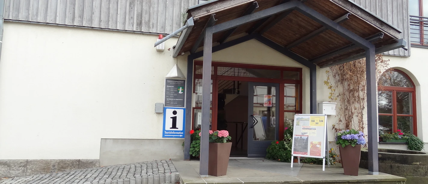Ingang van een toeristisch informatiecentrum met luifel, bloempotten en informatiebord; het gebouw heeft een lichtgekleurde gevel en houten bekleding.