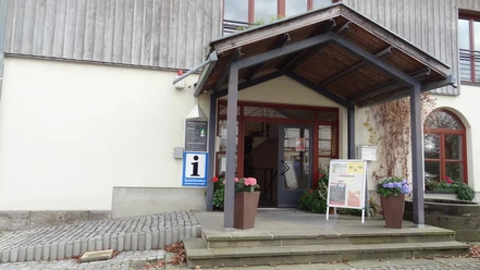 Touristinformation Gohrisch Eingang einer Touristinformation mit Überdachung, Blumenkübeln und Infotafel; das Gebäude hat eine helle Fassade und Holzverkleidung.